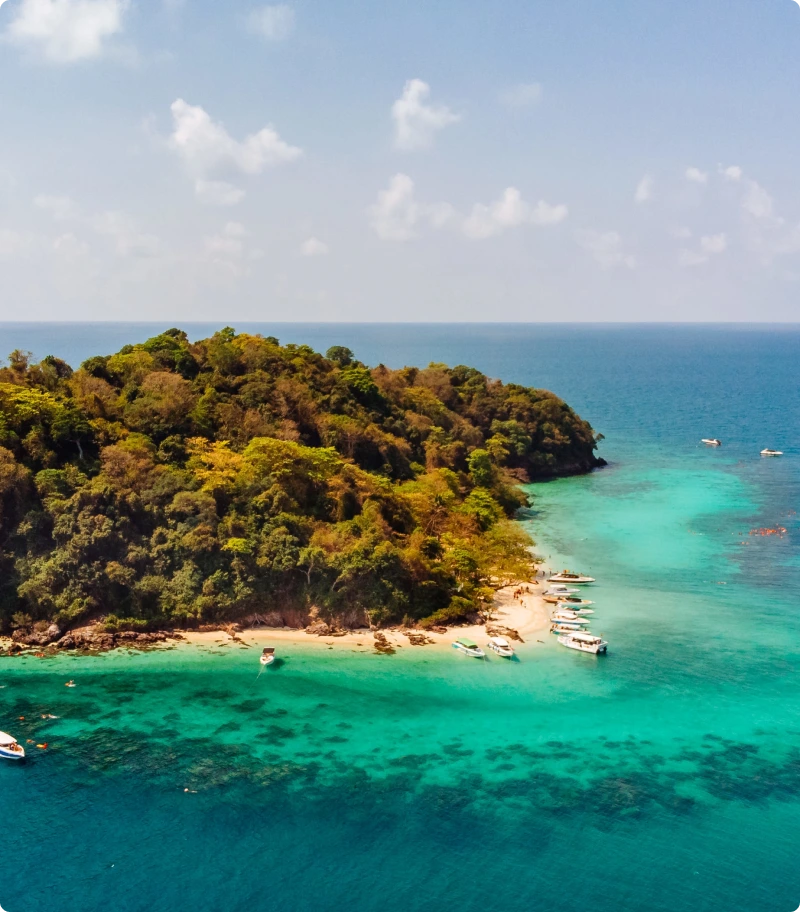 Island with lush greenery and clear blue water, with boats near the shore.